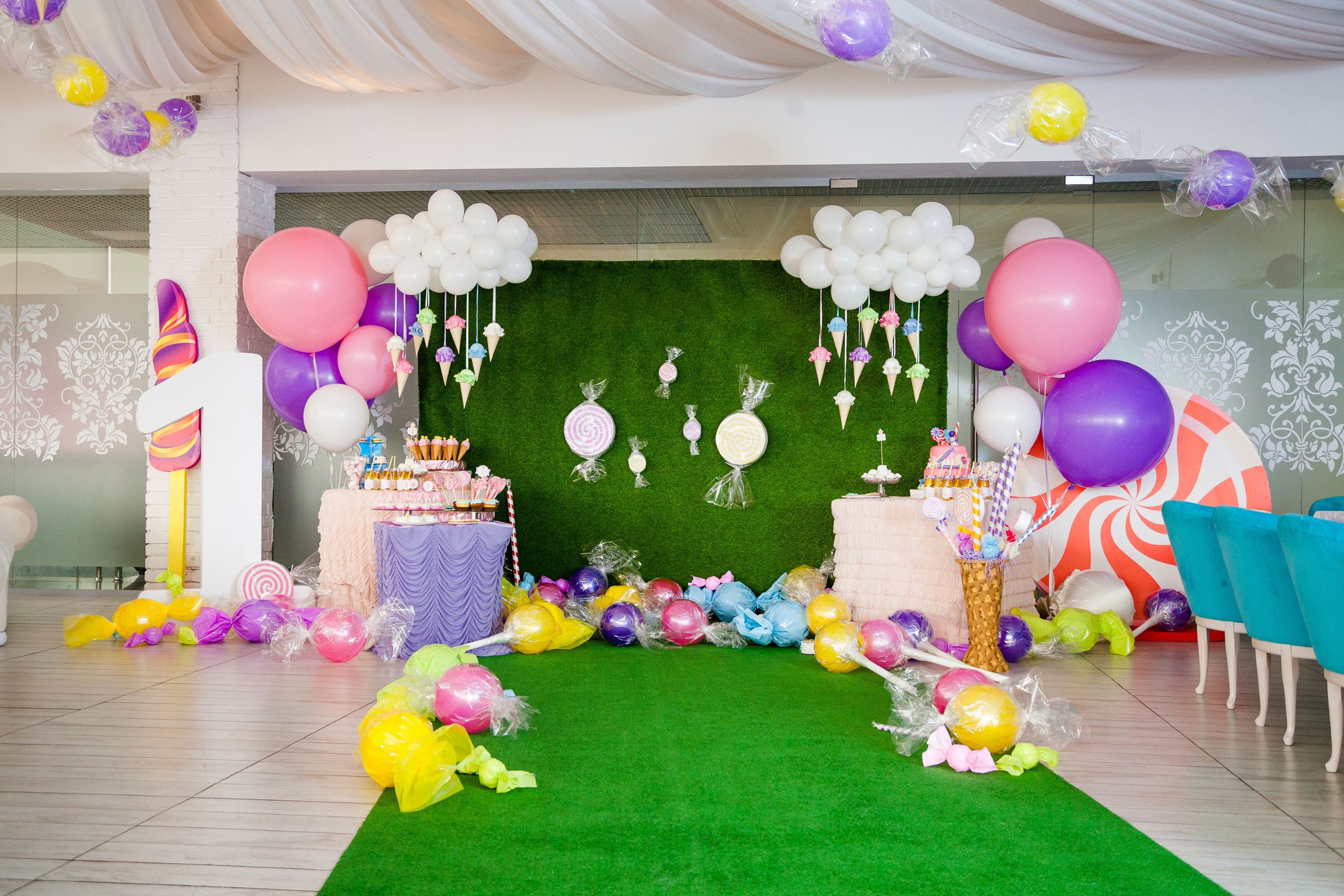 Auf dem Bild ist ein mit großen und kleinen weißen, rosanen und lila Luftballons geschmückter Raum zu sehen mit Dekoartikeln in Form von großen Bonbons, Zuckerstangen und Lutschern vor einem Tisch mit vielen Süßigkeiten.