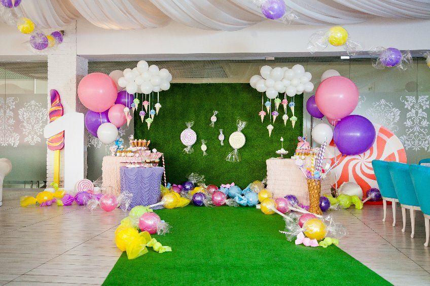 Auf dem Bild ist ein mit großen und kleinen weißen, rosanen und lila Luftballons geschmückter Raum zu sehen mit Dekoartikeln in Form von großen Bonbons, Zucherstangen und Lutschern vor einem Tisch mit vielen Süßigkeiten.