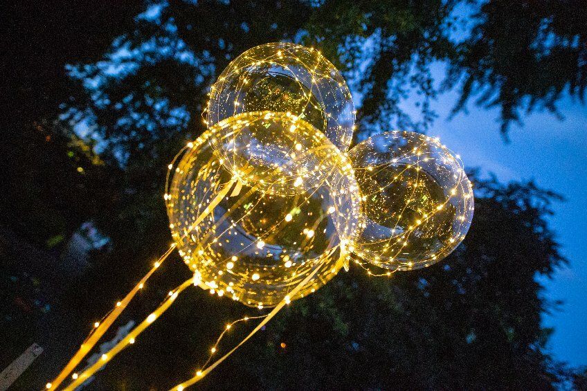 Auf den Bild sind in Dunkelheit schwebende transparente Luftballons, bestückt mit vielen LED-Lichtern, gehalten von LED-Lichterketten.