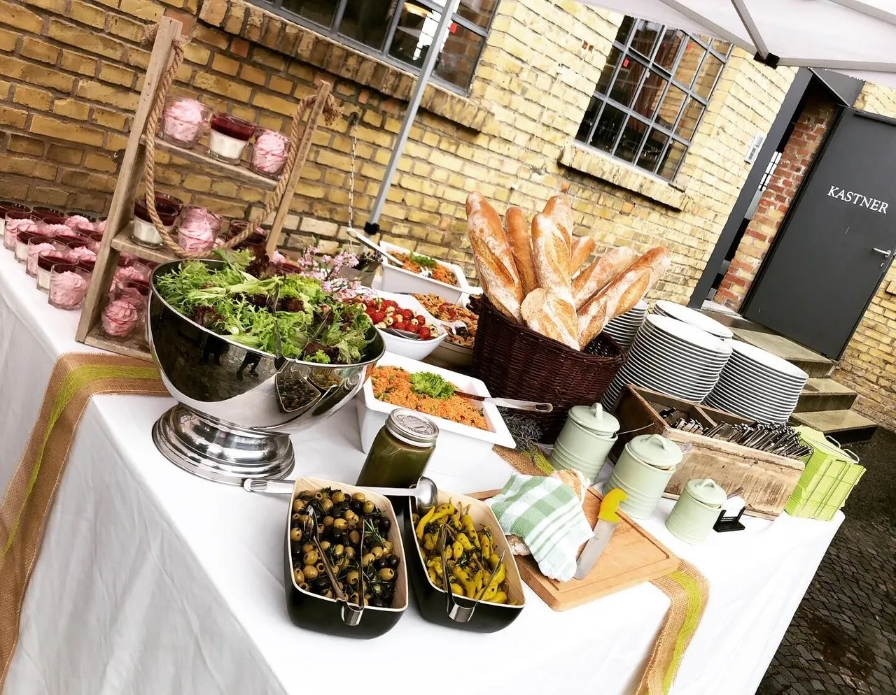 Auf dem Bild ist ein Büfett zu sehen, mit verschiedenen kalten Gerichten und Salaten sowie einen Korb voll Baguettes und im Hintergrund Gläser mit Nachtisch-Creme vor einer Hauswand