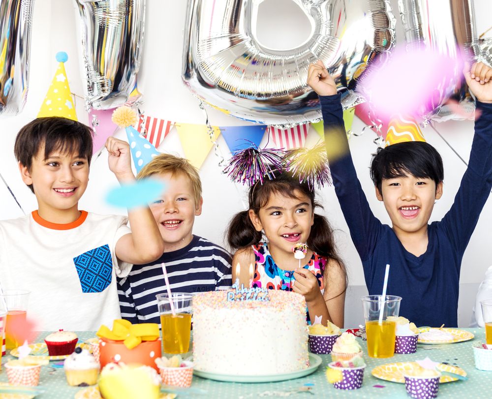 Zu sehen sind auf dem Bild drei Jungen und ein Mädchen, die sich richtig freuen, vor einem gedeckten Tisch auf dem eine große Geburtstagstorte steht