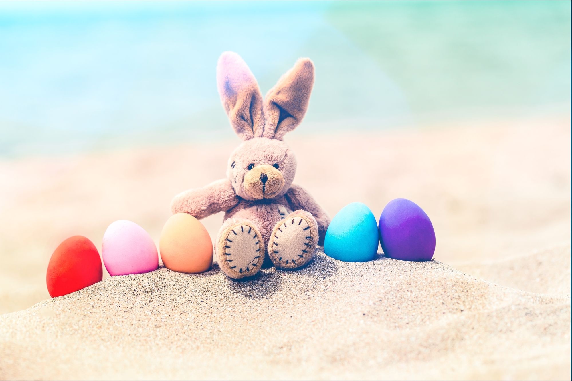 Das Bild zeigt zeigt eine Osterhasen-Stoffpuppe im Sand sitzend mit bunten Eiern neben sich.