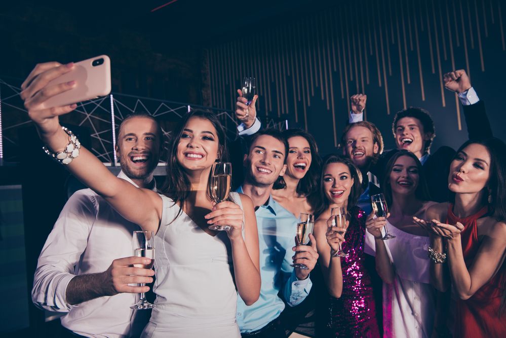 Auf dem Bild ist eine Gruppe junger Männer und Frauen mit Sektgläsern in der Hand zu sehen, die von einer jungen Frau im Vordergrund mit Ihrem Mobiltelefon in Selfieart fotografiert wird