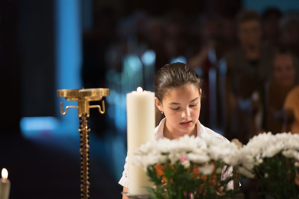 Auf dem Bild ist ein Mädchenkopf neben einer Kerze in einer Kirche zu sehen, die etwas vorliest in einem feierlichen Rahmen