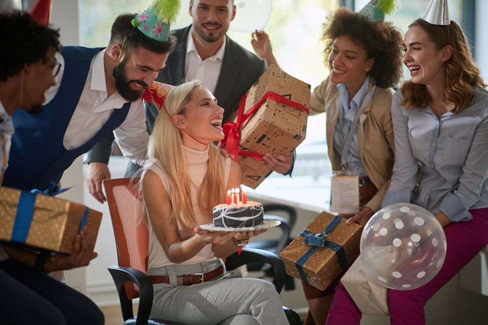 Auf dem Bild ist eine Partygesellschaft von jungen Männern und Frauen mit teils bunten Hütchen auf dem Kopf zu sehen und am Tisch sitzt das Geburtstagskind mit einer kleinen Torte in der Hand am Tisch und die Umstehenden halten Geschenke in der Hand
