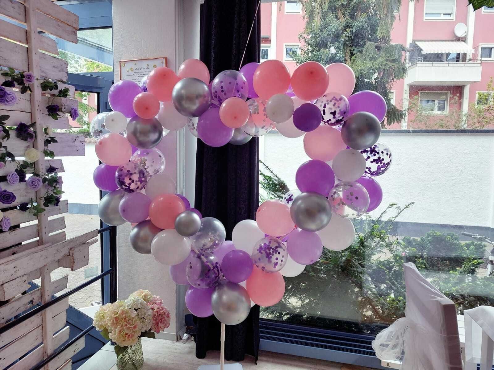 Das Bild zeigt einen großen herzförmigen Rahmen an dem viele Ballons in verschiedenen Farben befestigt sind