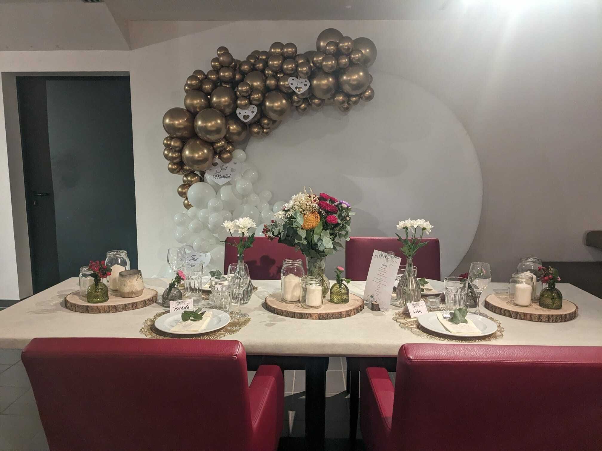 Auf dem Bild wird der Tisch eines Brautpaares gezeigt, festlich gedeckt mit Blumen und Kerzen und im Hintergrund hängt eine Ballongirlande mit goldglänzende und weißen Luftballons und einem Hinweistext "Just married". 