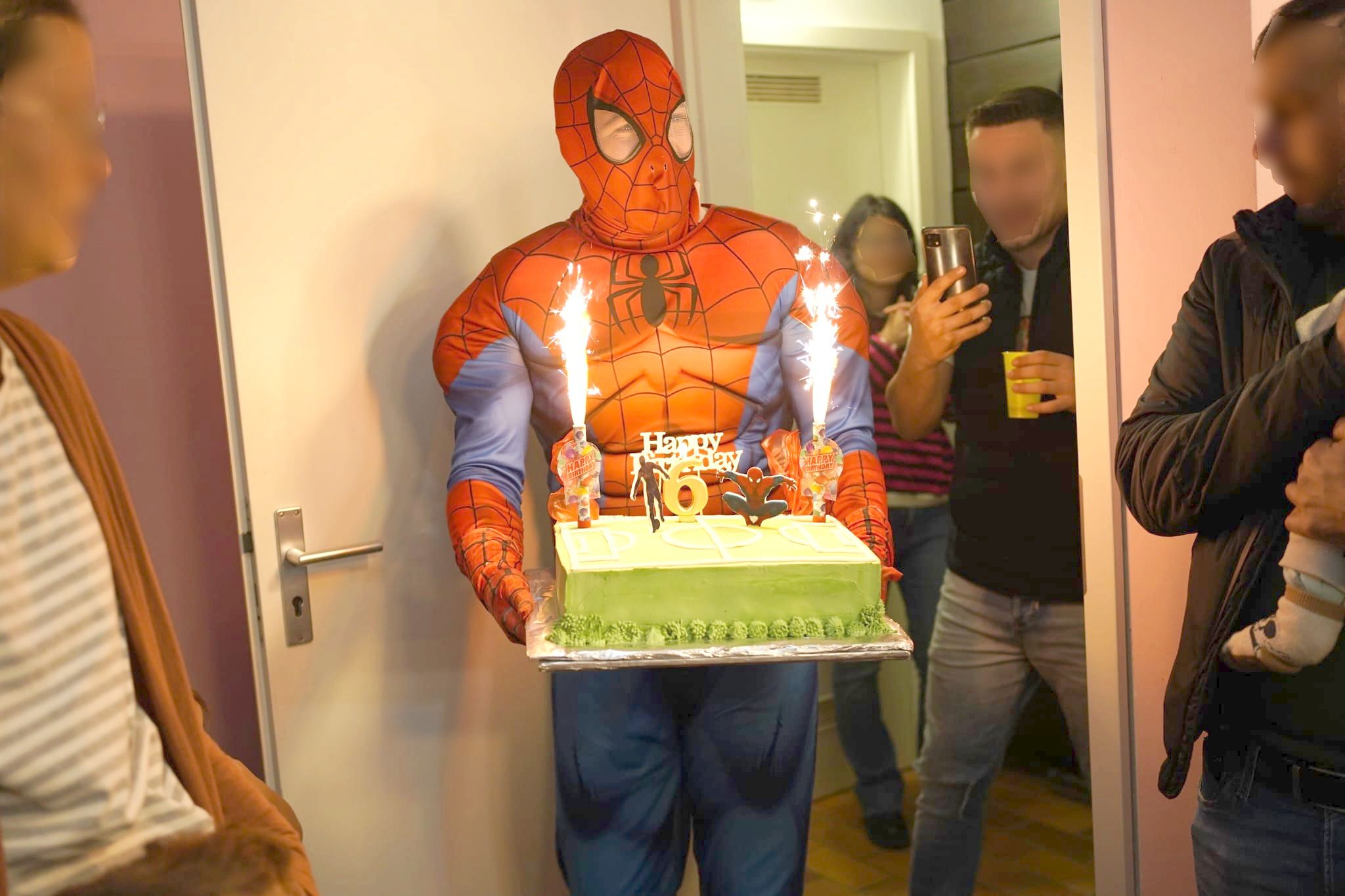 Auf dem Bild ist ein Animateur als Spiderman verkleidet, der mit einer Torte in den Händen gerade ins Zimmer tritt. Im Hintergrund sind Gäste zu sehen, die den Event fotografieren mit ihrem Mobiltelefon..