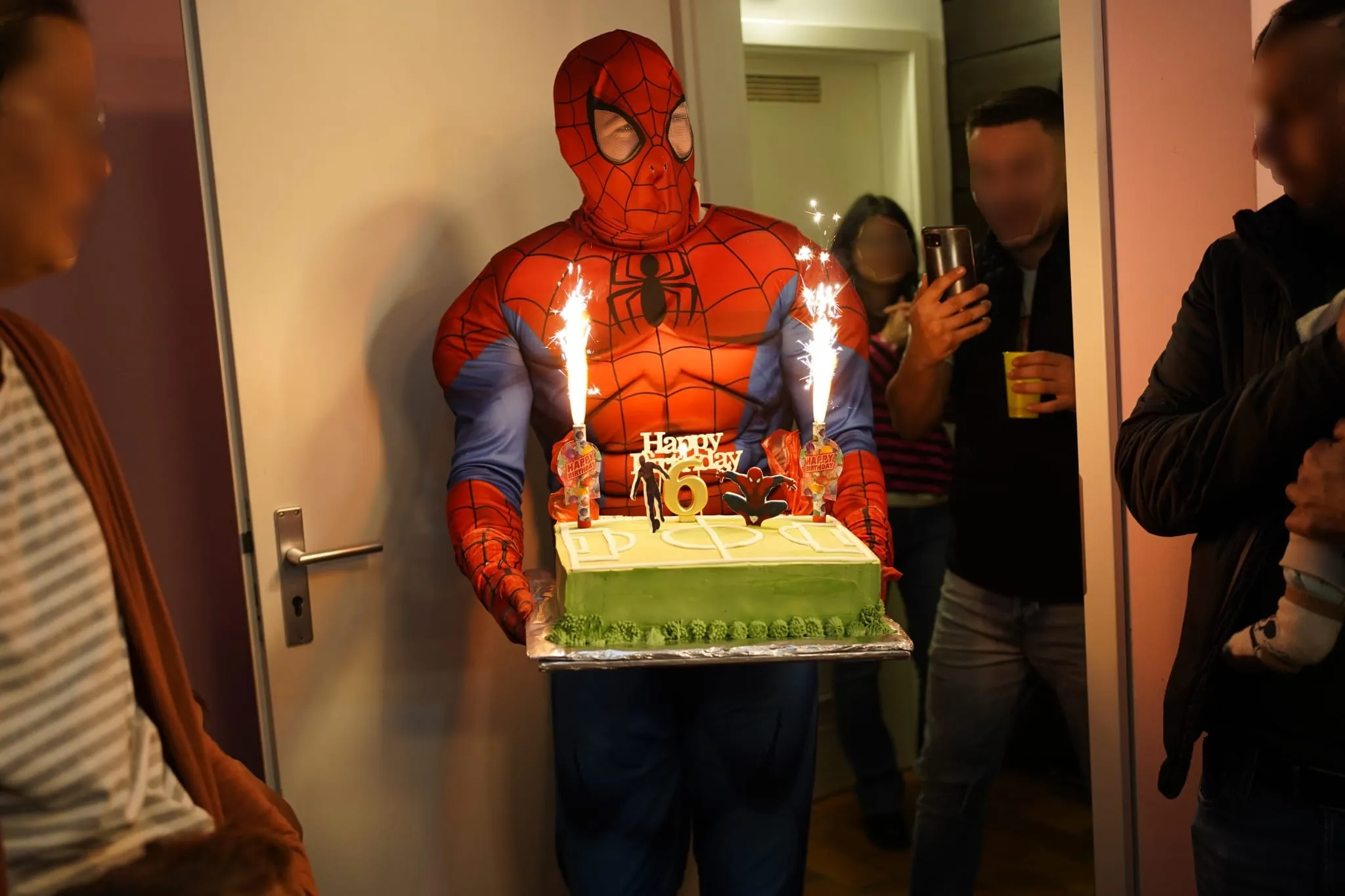 Auf dem Bild ist ein Animatuer als Spiderman verkleidet, der mit einer Torte in den Händen gerade ins Zimmertritt. Im Hintergrund sind Gäste zu sehen, die den Event fotografieren mit ihrem Mobilphone.